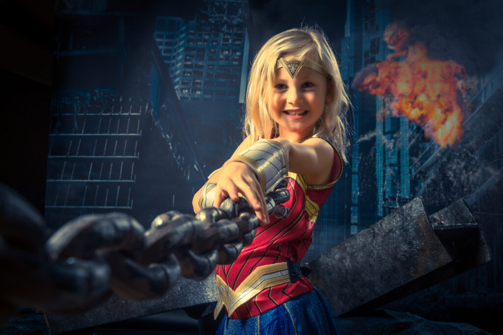 Young girl smiling while holding onto a metal chain, dressed in a colourful superhero costume during a themed studio photoshoot.